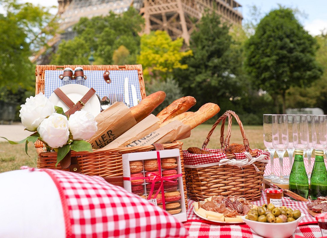 Picnic setup with a blanket, basket, and food on the grass near the Eiffel Tower on a sunny day.